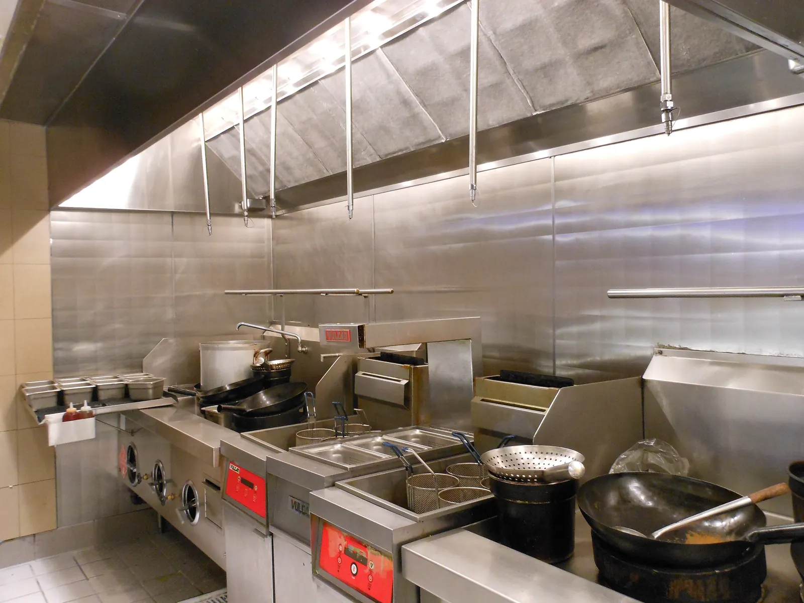 Stainless-steel exhaust hood with baffle filters running above a commercial cooking line with fryers and a wok range